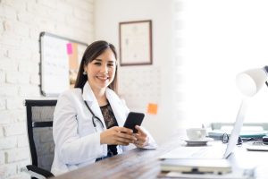 Minha clínica nas redes sociais: devo ou não aderir? 1 Médica sorridente usando celular em ambiente de clínica médica