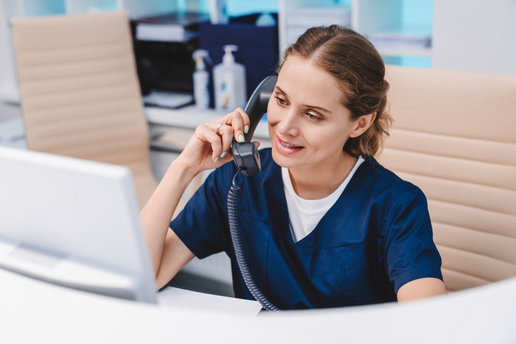 Jovem recepcionista em clínica falando ao telefone enquanto utiliza computador