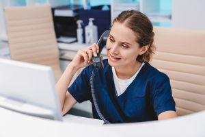 Jovem recepcionista em clínica falando ao telefone enquanto utiliza computador
