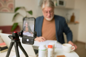 Qual é a importância de um bom marketing para médicos? 1 Médico em frente a um computador usando o seu celular para gravar vídeos como estratégia de marketing digital