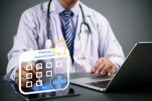 Médico sentado em seu consultório usando o seu notebook e tablet para marcar consultas