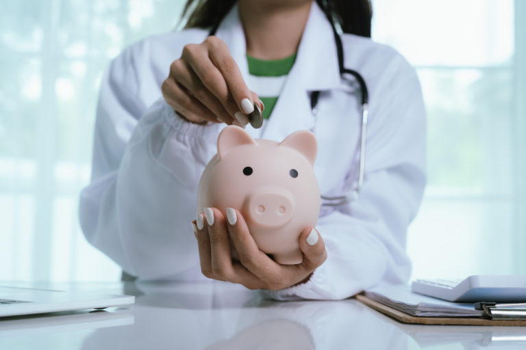 Médica guardando uma moeda dentro de um cofre de porquinho que está em sua outra mão